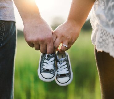 Woman And Man Holding Black Crib Shoes Standing Near Green Grass