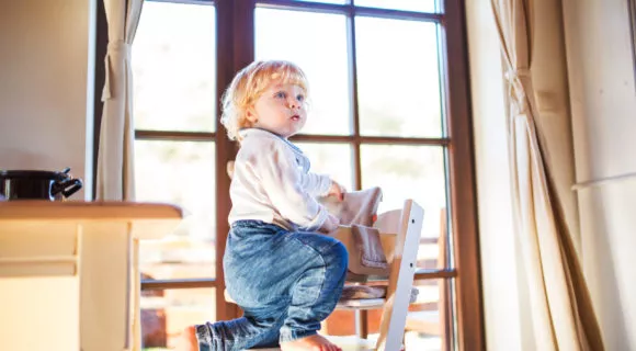 Little toddler boy climbing on wooden highchair at home. Domestic accident. Dangerous situation at home.